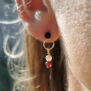 Peut inclure: Boucle d'oreille dorée avec une perle noire, un anneau doré, une perle blanche et une perle rouge. La boucle d'oreille est suspendue au lobe de l'oreille. Un ear cuff doré est également visible.
