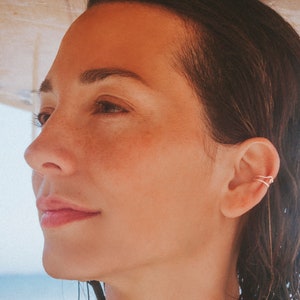 Könnte beinhalten: Eine Frau mit dunklem Haar und hellem Teint, die einen goldenen Ohrklemmen trägt. Sie hält ein Surfbrett über sich. Der Hintergrund zeigt einen Strand und das Meer.