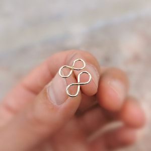 Op de afbeelding: Een gouden oneindigheidssymbool bedel, gemaakt van draad, wordt in een hand gehouden. De bedel is een dubbel oneindigheidssymbool, met twee lussen die met elkaar verweven zijn.