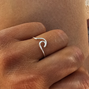 Può includere: Un anello d'argento a forma di onda. L'anello è fatto di un sottile pezzo di metallo curvo che ha la forma di un'onda. L'anello viene indossato su un dito.