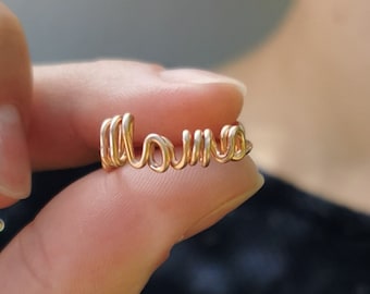 Könnte beinhalten: Ein goldfarbener Drahtring mit dem Wort "Mama" in Schreibschrift. Der Ring wird zwischen zwei Fingern vor einem verschwommenen Hintergrund gehalten. Der Ring besteht aus einem dünnen, glänzenden Draht.