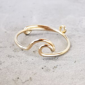May include: A gold-toned ring with a wave design. The ring is made of wire and has a small loop at the top.