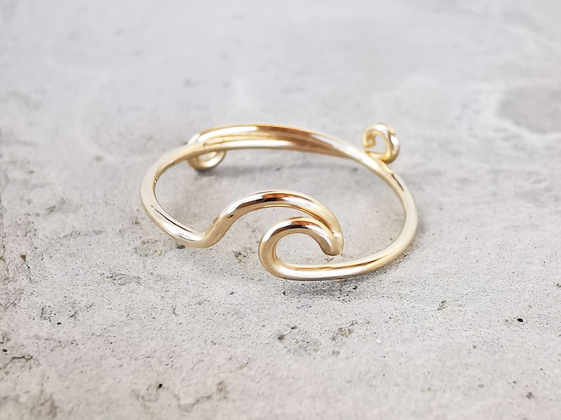 May include: A gold-toned ring with a wave design. The ring is made of wire and has a small loop at the top.