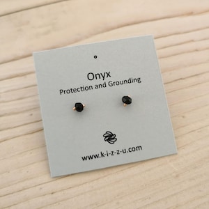 Puede incluir: Un par de pendientes de botón de ónix negro sobre una tarjeta gris claro. La tarjeta dice "Onyx Protección y Enraizamiento" con una dirección web. Los pendientes están engarzados en metal dorado.