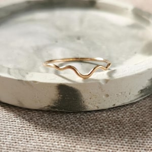 Può includere: Un sottile anello in oro caratterizzato da un delicato design ondulato o sinuoso continuo lungo la metà superiore della fascia. Il metallo presenta una finitura lucida e riflettente che cattura la luce, creando una silhouette liscia e fluida.