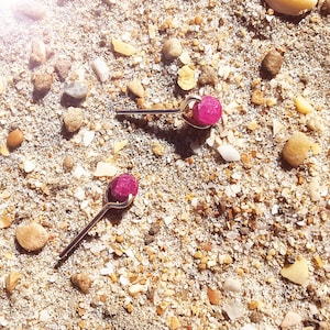 Könnte beinhalten: Zwei silberfarbene Ohrstecker mit hellrosa, abgerundeten Edelstein-Akzenten. Die Ohrringe liegen auf einem Hintergrund aus Sand und kleinen Kieselsteinen, was eine natürliche, strandähnliche Ästhetik erzeugt. Die Ohrringe sind schlicht und elegant.