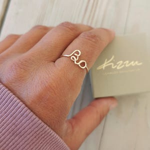 Può includere: Un anello in filo d'argento con la parola "Bo" indossato su un dito. L'anello ha un design minimalista. Una scatola colorata chiara con la parola "kizzu" è sullo sfondo.