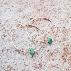 Puede incluir: Un par de pendientes de aro de oro rosa, cada uno adornado con una pequeña piedra preciosa verde facetada. Los pendientes se exhiben sobre una superficie texturizada de tono neutro, resaltando su delicado diseño y color.