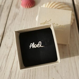 Peut inclure: Une bague en argent avec le nom "Abel" écrit en cursive, présentée dans un écrin ouvert à intérieur noir. La boîte est posée sur une surface en bois clair, avec un coquillage et une boîte fermée en arrière-plan.