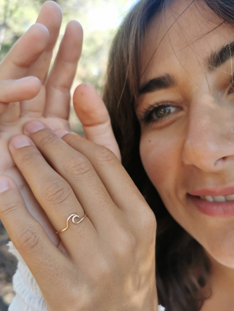 May include: A delicate gold ring featuring a simple wave design. The ring is worn on the finger of a person with brown hair.