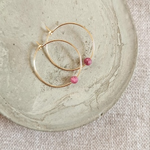 Op de afbeelding: Een paar gouden oorringen met een klein roze edelsteentje aan elke ring.