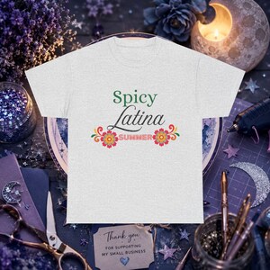 May include: A light gray short-sleeve t-shirt featuring the green text Spicy, the black cursive text Latina, and the red text SUMMER. The design includes two colorful floral accents with red, orange, and yellow petals on either side of the word SUMMER.