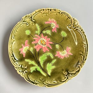 May include: A round ceramic plate featuring a glossy olive green glaze with a decorative embossed border. The center displays a hand-painted illustration of pink flowers with yellow centers and green leaves, accented by fine black outlines.