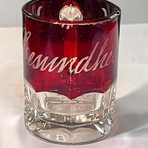 May include: A small cylindrical shot glass featuring a deep red upper section and a clear glass base. The red portion is etched with the white cursive text "Gesundheit". The glass has a thick, sturdy bottom and a smooth, polished rim.