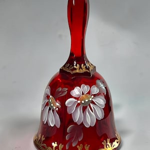 May include: A ruby red glass bell featuring hand-painted white daisy flowers with yellow centers. The bell is decorated with gold filigree patterns along the top neck and the bottom rim, providing a contrast to the deep red translucent glass body.