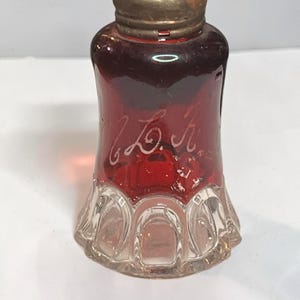May include: A vintage salt or pepper shaker featuring a deep red glass body with an etched script monogram on the front. The base is made of clear glass with a molded arch pattern, and the top is a tarnished metal cap with small circular holes.