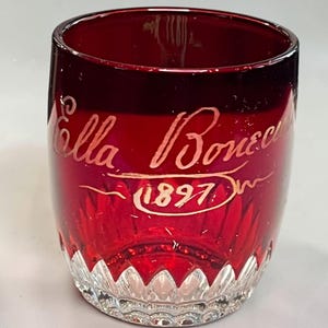 May include: A deep red glass tumbler featuring a clear, faceted base with a scalloped edge. The exterior surface is hand-painted with gold-coloured script text that reads "Ella Bonec" above the year "1897" enclosed in an oval line design.
