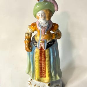 May include: A porcelain figurine of a person wearing a wide-brimmed green hat with a feather, a yellow and blue bodice, and a long blue skirt with a central yellow panel. The figure features gold-painted accents and stands on a round base.
