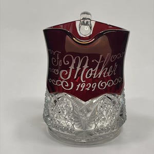 May include: A clear and ruby red glass pitcher featuring a cut crystal pattern on the lower half. The upper red section is etched with the text To Mother and the year 1929 in a decorative script font. The pitcher has a clear glass handle.