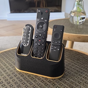 Puede incluir: Un soporte para mandos a distancia en negro y dorado con tres ranuras verticales, cada una con un mando negro. El soporte tiene un exterior negro texturizado y un revestimiento interior dorado liso, diseñado para organizar dispositivos.