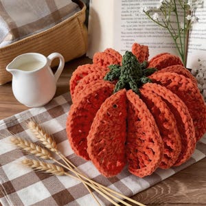 Puede incluir: Una calabaza de ganchillo con segmentos de hilo naranja brillante y un tallo verde oscuro. La calabaza se muestra junto a una jarra de cer&aacute;mica blanca, tallos de trigo secos y un libro con texto visible, sobre un pa&ntilde;o a cuadros marr&oacute;n y blanco.