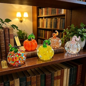 Puede incluir: Cuatro calabazas decorativas de vidrio dispuestas en un estante de madera. Una presenta un patrón de confeti multicolor, una es naranja sólido, una es amarilla brillante y una es transparente con un tallo rosa. Cada una tiene un tallo curvo.