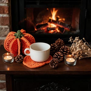 Puede incluir: Una taza de cer&aacute;mica blanca sobre un posavasos de ganchillo naranja con forma de calabaza junto a una calabaza de ganchillo m&aacute;s grande. El conjunto incluye pi&ntilde;as marrones, flores secas y dos portavelas de cristal con velas encendidas.