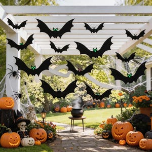 May include: A collection of black hanging bat decorations with glowing green eyes, suspended from a white pergola. The scene includes carved jack-o'-lantern pumpkins, a skeleton figure, a black cauldron on a stand, and artificial spider webs.