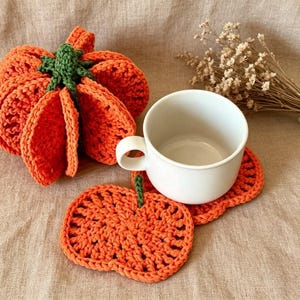 May include: A bright orange crocheted pumpkin with a green stem, a white ceramic mug, and two flat orange crocheted pumpkin-shaped coasters. The coasters are textured with a grid-like pattern and feature small green stems at the top.