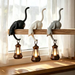 Puede incluir: Tres figuras con forma de gato en negro, blanco y gris, cada una posada sobre un estante de madera. Cada gato tiene una cola larga que cuelga hacia abajo, sosteniendo una pequeña linterna de color bronce con una luz brillante en su interior.