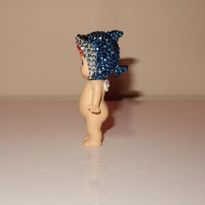 K&ouml;nnte beinhalten: Kleine Figur einer nackten, baby&auml;hnlichen Figur mit einem blau-wei&szlig;en Haifischflossen-Hut. Der Hut ist mit glitzernden blauen Strasssteinen bedeckt. Die Figur steht auf einer wei&szlig;en Oberfl&auml;che.