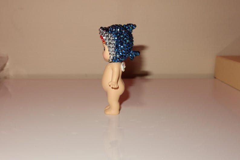 K&ouml;nnte beinhalten: Kleine Figur einer nackten, baby&auml;hnlichen Figur mit einem blau-wei&szlig;en Haifischflossen-Hut. Der Hut ist mit glitzernden blauen Strasssteinen bedeckt. Die Figur steht auf einer wei&szlig;en Oberfl&auml;che.