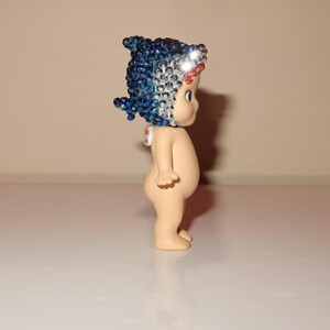 K&ouml;nnte beinhalten: Kleine Figur eines nackten Babys mit blau-silbernem Schmuckkopfschmuck. Die Figur steht auf einer wei&szlig;en Oberfl&auml;che. Das Baby hat einen hellen Hautton und ist zur Seite gedreht.