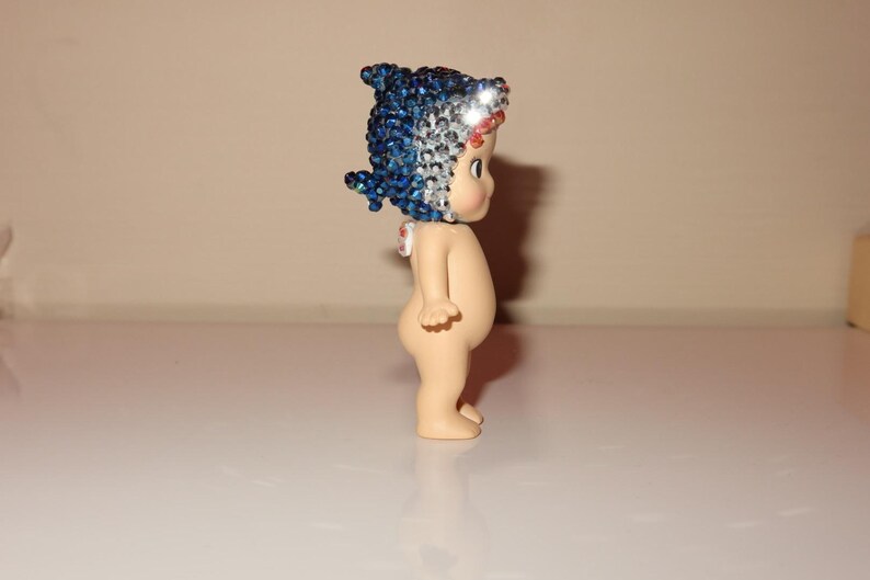 K&ouml;nnte beinhalten: Kleine Figur eines nackten Babys mit blau-silbernem Schmuckkopfschmuck. Die Figur steht auf einer wei&szlig;en Oberfl&auml;che. Das Baby hat einen hellen Hautton und ist zur Seite gedreht.