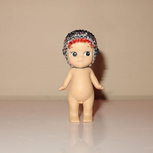 K&ouml;nnte beinhalten: Kleine, nacktfarbene Figur einer baby&auml;hnlichen Figur mit einer detaillierten, mit Perlen besetzten M&uuml;tze. Die M&uuml;tze hat ein Muster aus schwarzen, silbernen und roten Perlen. Die Figur hat gro&szlig;e, ausdrucksstarke Augen und steht auf einer wei&szlig;en Oberfl&auml;che.