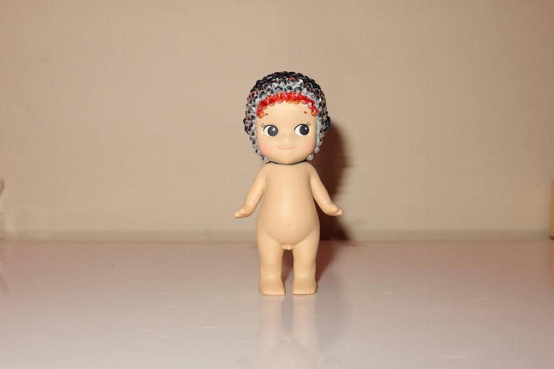 K&ouml;nnte beinhalten: Kleine, nacktfarbene Figur einer baby&auml;hnlichen Figur mit einer detaillierten, mit Perlen besetzten M&uuml;tze. Die M&uuml;tze hat ein Muster aus schwarzen, silbernen und roten Perlen. Die Figur hat gro&szlig;e, ausdrucksstarke Augen und steht auf einer wei&szlig;en Oberfl&auml;che.