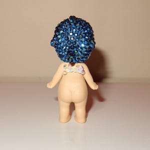 K&ouml;nnte beinhalten: Kleine Figur mit blauem, geschm&uuml;cktem Kopf und nacktem K&ouml;rper. Die Figur hat ein kleines, dekoratives St&uuml;ck auf dem R&uuml;cken. Die Figur steht auf einer wei&szlig;en Oberfl&auml;che.