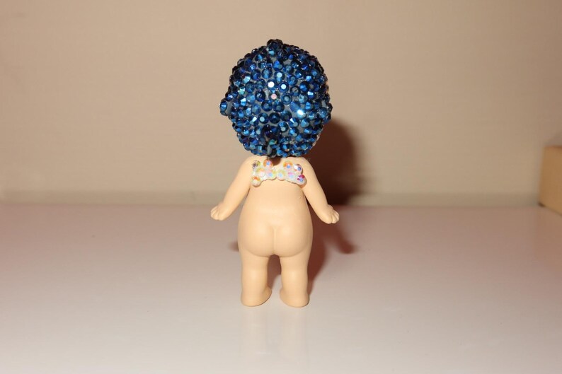 K&ouml;nnte beinhalten: Kleine Figur mit blauem, geschm&uuml;cktem Kopf und nacktem K&ouml;rper. Die Figur hat ein kleines, dekoratives St&uuml;ck auf dem R&uuml;cken. Die Figur steht auf einer wei&szlig;en Oberfl&auml;che.