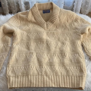 Könnte beinhalten: Ein hellbeige Langarmpullover mit V-Ausschnitt. Der Pullover hat ein strukturiertes Strickmuster mit geometrischen Formen sowie gerippte Bündchen und Saum. Ein kleines blaues Etikett ist am Kragen sichtbar.