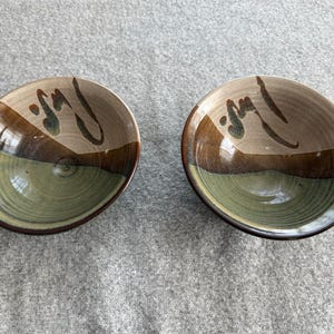 May include: Two round ceramic bowls featuring a tri-color glaze design. The interior of each bowl displays a light beige upper section, a dark brown middle band, and a sage green base, accented with dark brown brushstroke markings on the rim.