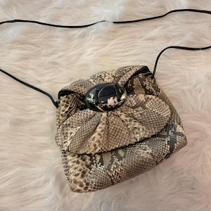 May include: A crossbody bag featuring a snakeskin pattern in shades of beige, gray, and black. The bag has a gathered, rounded flap closure secured by a large, smooth, oval-shaped black stone ornament and includes a thin black shoulder strap.