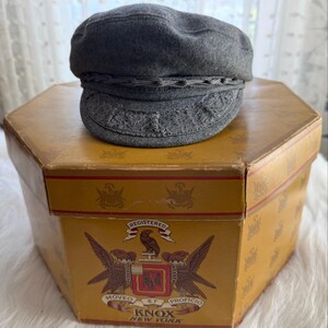 May include: A gray wool cap with a decorative band sits atop a vintage hexagonal box. The cap has a rounded crown and a detailed brim. The box is yellow with a printed design and the text "KNOX NEW YORK".