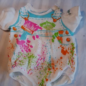 May include: A white short-sleeved bodysuit featuring a printed graphic of blue overalls with orange buttons. The design includes a paintbrush, paint splatters in pink, green, and orange, and the text Cabbage Patch Kids 1984 OAA Inc.