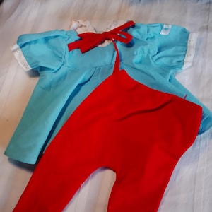 Op de afbeelding: Een lichtblauw shirt met korte mouwen, een witte kraag van broderie anglaise en afgewerkt met kanten randen aan de mouwen. Bij de hals zit een felrode stoffen strik. Het setje wordt gecombineerd met een effen rode legging voor een kleurrijk geheel.