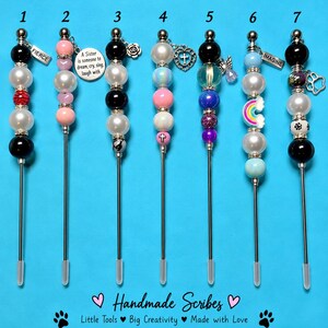 May include: Seven metal scribe tools with decorative beads and charms. Each tool features a unique arrangement of colored beads, including black, white, pink, and blue, with charms like a cross, heart, paw print, and text tags reading FIERCE and IMAGINE.