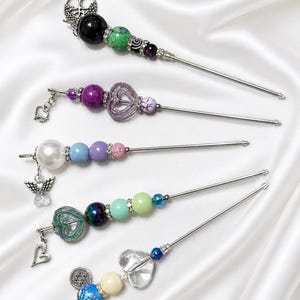 May include: Five metal hair sticks decorated with assorted beads and charms. The sticks feature various designs including a tree of life, hearts, an angel, and a Star of David, with beads in shades of black, green, purple, blue, pink, and white.