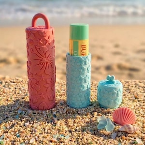 May include: Three decorative containers and a small trinket box are displayed on a sandy beach. The containers are coral, light blue, and teal, with floral and seashell patterns. A tube of lip balm is in one container. A seashell and a small flower are also present.