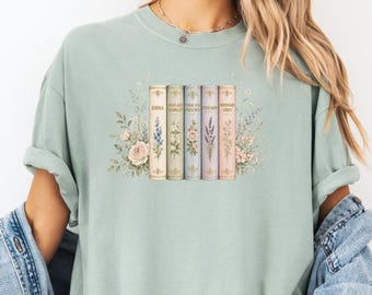 Pode incluir: Uma t-shirt de gola redonda em verde sálvia com um gráfico de cinco lombadas de livros de estilo vintage alinhadas, cada uma decorada com ilustrações florais delicadas e texto, rodeadas por um grupo de flores botânicas de tons suaves.