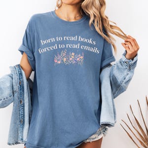 May include: A blue short-sleeved t-shirt featuring the white text "born to read books forced to read emails" printed above a colorful graphic of various wildflowers in shades of pink, orange, purple, and yellow on the center chest.