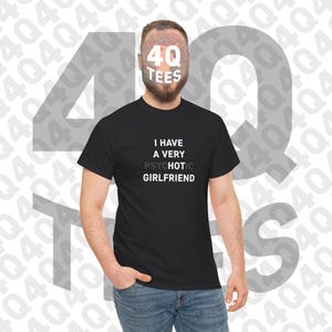 May include: A black short-sleeved t-shirt featuring white text that reads I HAVE A VERY PSYCHOTIC HOT GIRLFRIEND, with the word PSYCHOTIC partially obscured by a greyed-out effect. The shirt is worn with blue denim jeans.